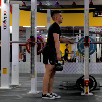 Jaskółka z kettlebell — pozycja startowa: stanie jednonóż, kettlebell w rękach, asekuracja o sztangę